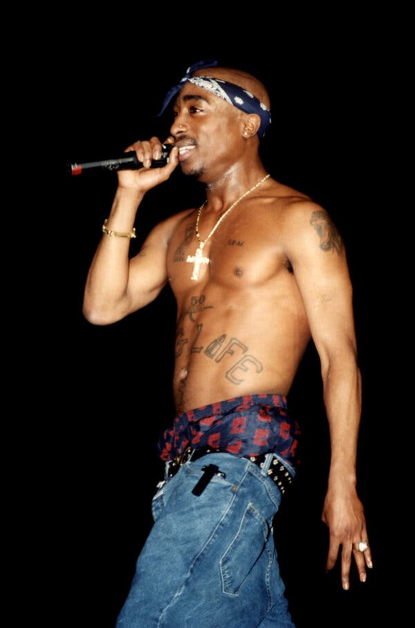 A bare-chested Tupac Shakur holds a microphone to his mouth while wearing a blue bandanna around his head, a gold cross and low jeans.