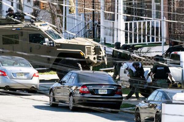 Heavily armed police officers dressed in black surround a house as an armored vehicle is parked in front. 