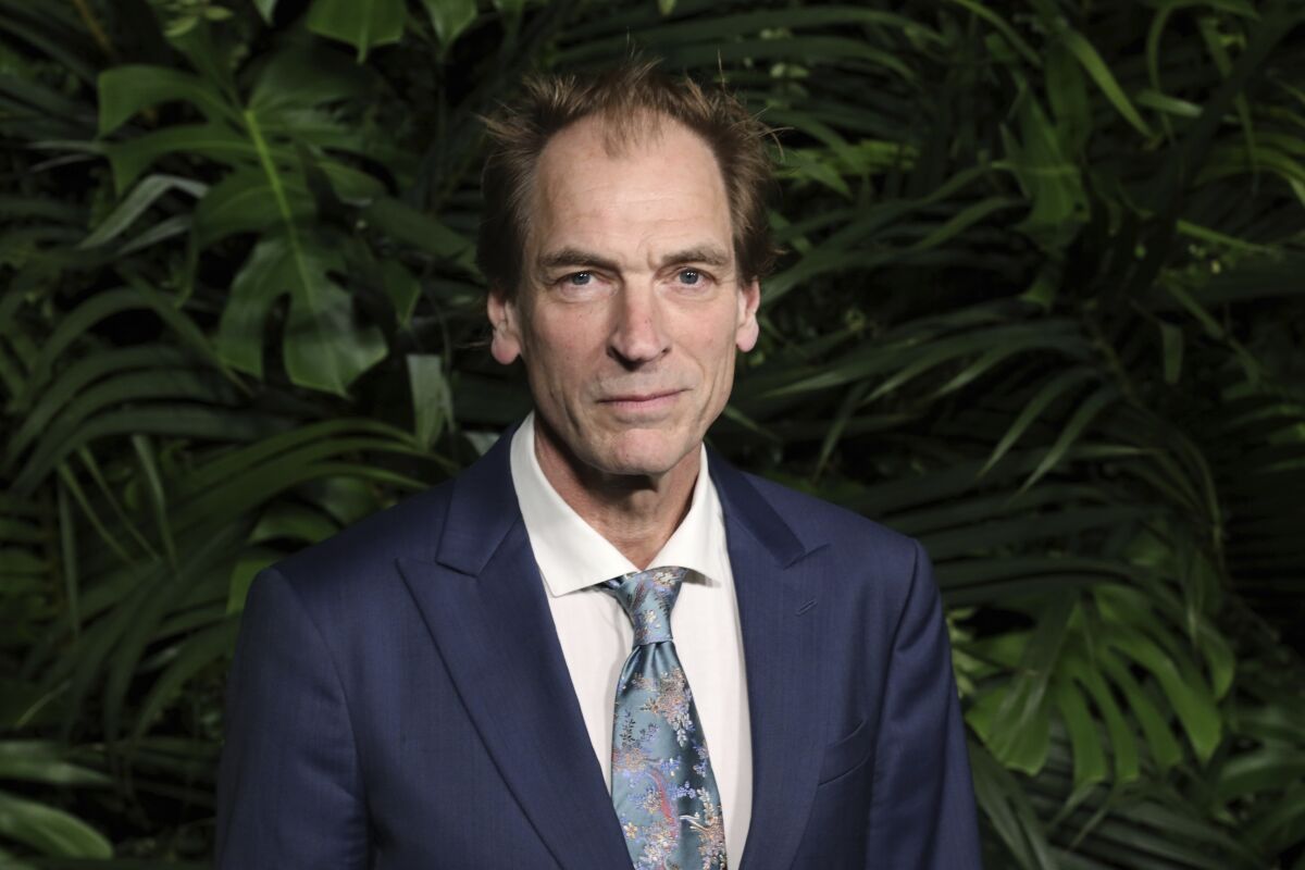 A middle-aged man in a suit and tie poses in front of a leafy green backdrop