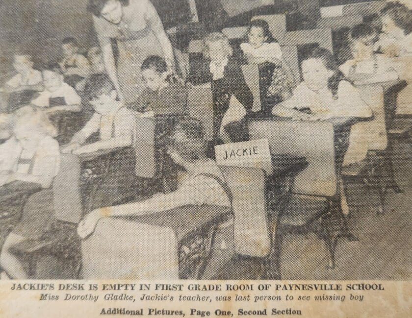 A black and white newspaper clipping of a photo of first grade students sitting at there desks as a teacher leans over one of the students to help with their work. The second desk back from the right is empty. 