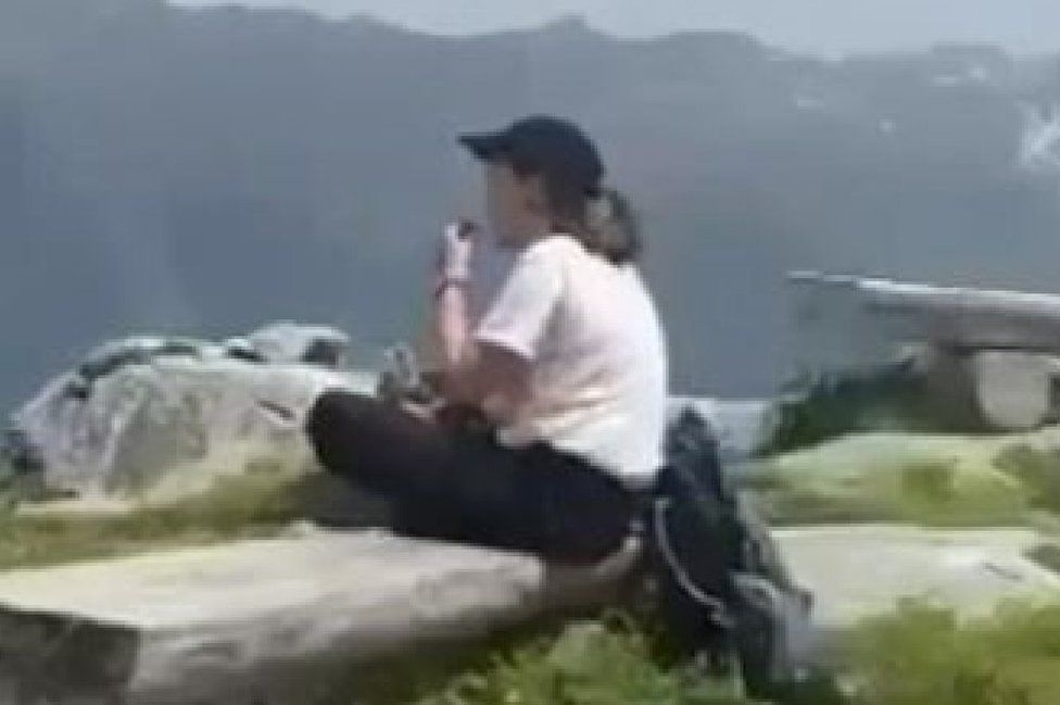A still of a woman in a white shirt and black cap sitting on a rock eating