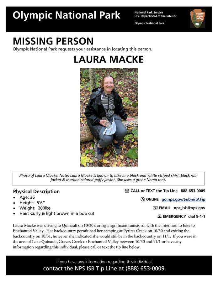 Laura Macke sits on a log in a forested area smiling at the camera. She is wearing a black jacket, black and white striped shirt, gray pants and gray shoes. She has light skin, brown hair that is pulled back and wears glasses.